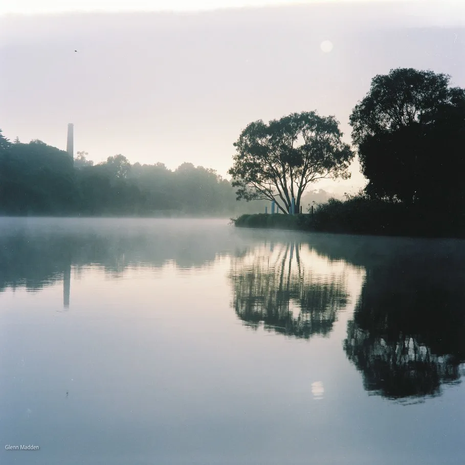 Misty dawn on the Barwon River with gum trees and an old industrial chimney reflected in perfectly still water, a hazy full moon visible in the pale pastel sky