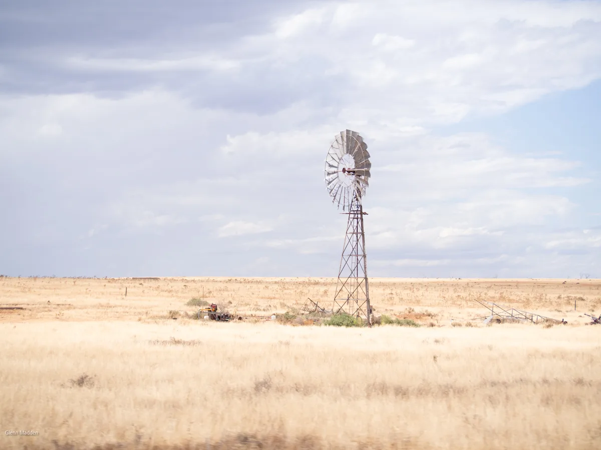 A solitary vintage farm windmill standing tall in flat sun-bleached grassland under a dramatic cloud-streaked sky in outback Victoria, with a pump at its base