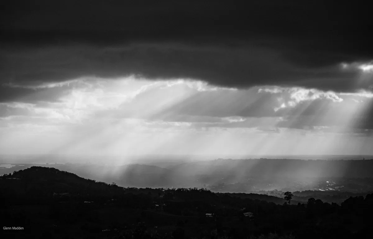 Black-and-white panorama of dramatic crepuscular rays bursting through storm clouds over the Toowoomba escarpment, mist hanging across the valley below