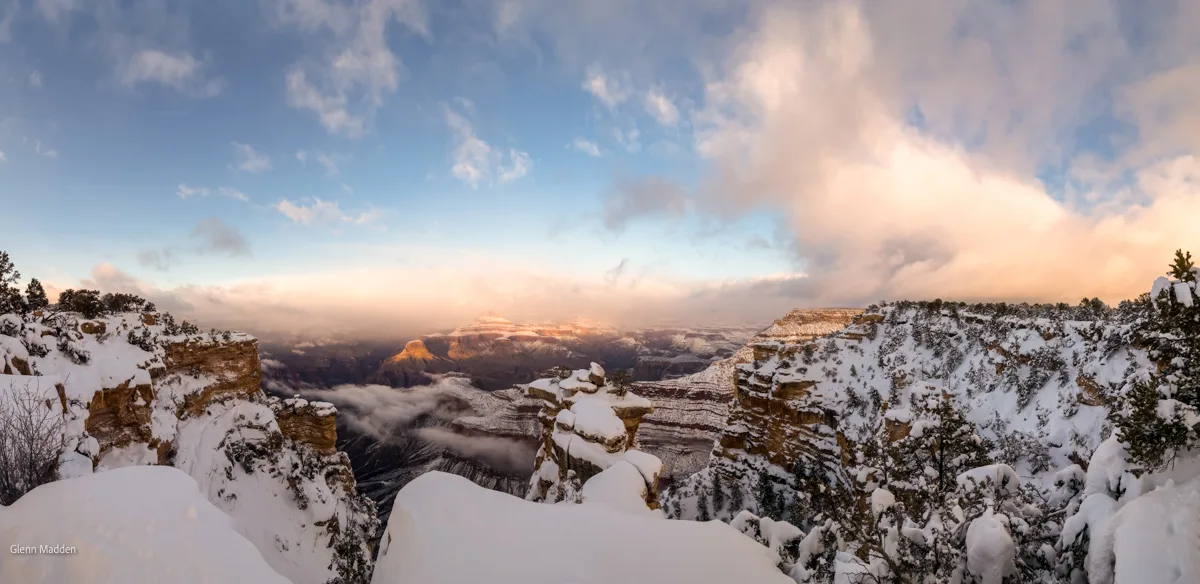 Panoramic winter view from the South Rim of the Grand Canyon after heavy snowfall, snow-dusted canyon walls glowing in warm sunrise light, low cloud filling the gorge depths