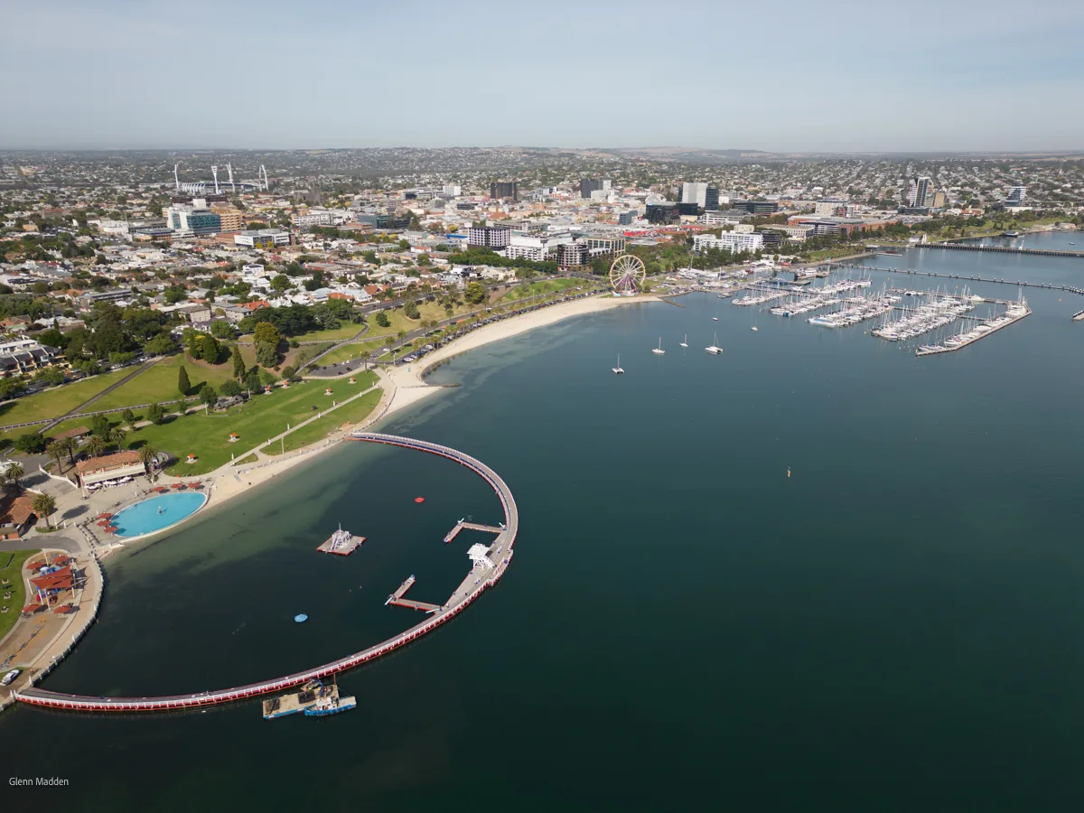 Aerial view of Geelong's Eastern Beach foreshore featuring the iconic curved heritage swimming enclosure, a packed sailing marina, Ferris wheel, and the city skyline stretching inland