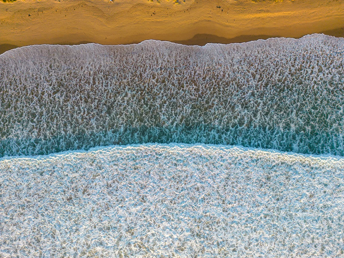 Aerial top-down abstract of a frothy wave washing across golden sand at Lorne beach, white sea foam creating intricate textures against pale turquoise water