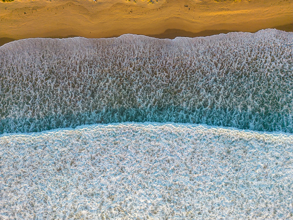 Aerial top-down abstract of a frothy wave washing across golden sand at Lorne beach, white sea foam creating intricate textures against pale turquoise water