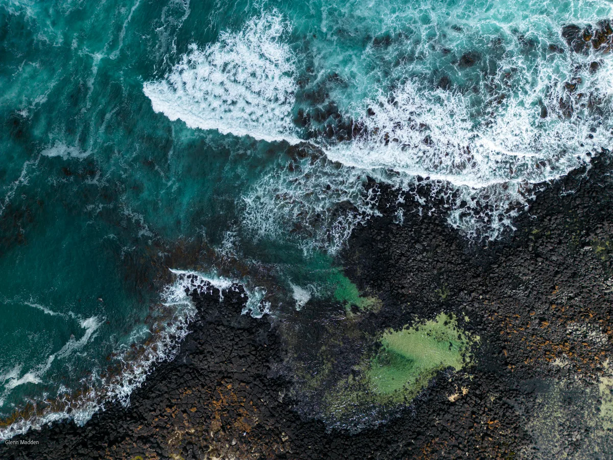Aerial abstract of turquoise ocean waves crashing over dark volcanic basalt rock platform at Port Fairy, with vivid green algae pooled in the rock crevices