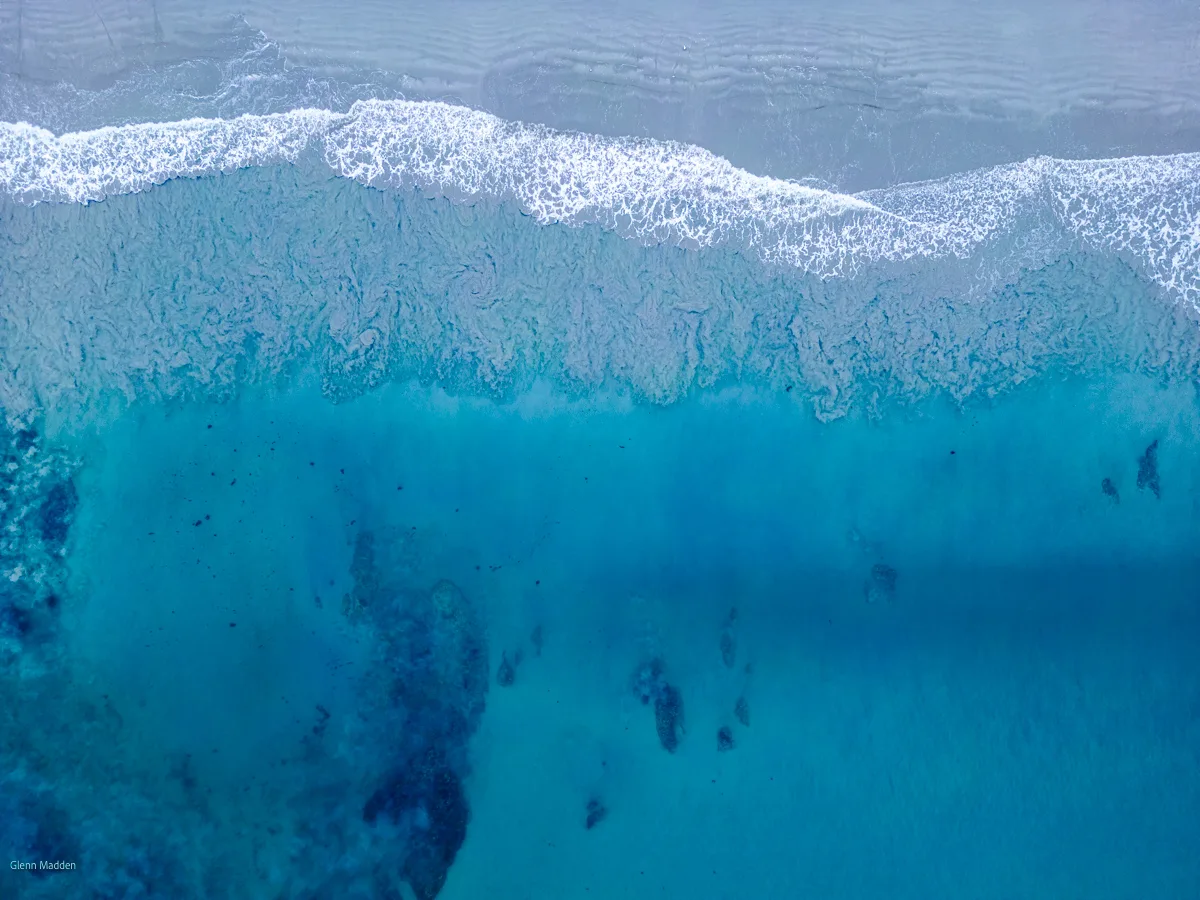 Aerial top-down view of a breaking wave along Portland's white sand beach with vivid aquamarine shallow water, dark reef shadows visible beneath the surface