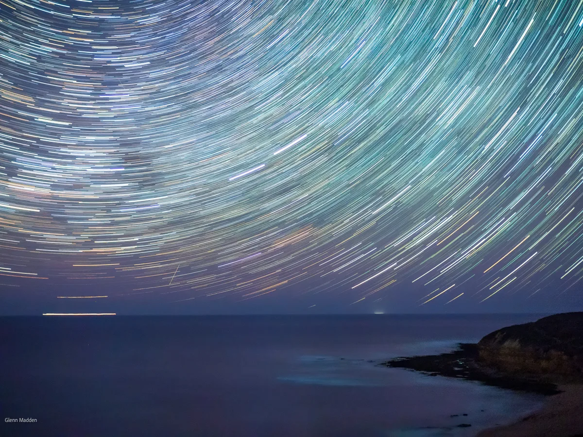 Long-exposure star trails forming sweeping concentric arcs across the night sky over a calm ocean horizon at Bells Beach, rocky coastal cliffs silhouetted in the foreground